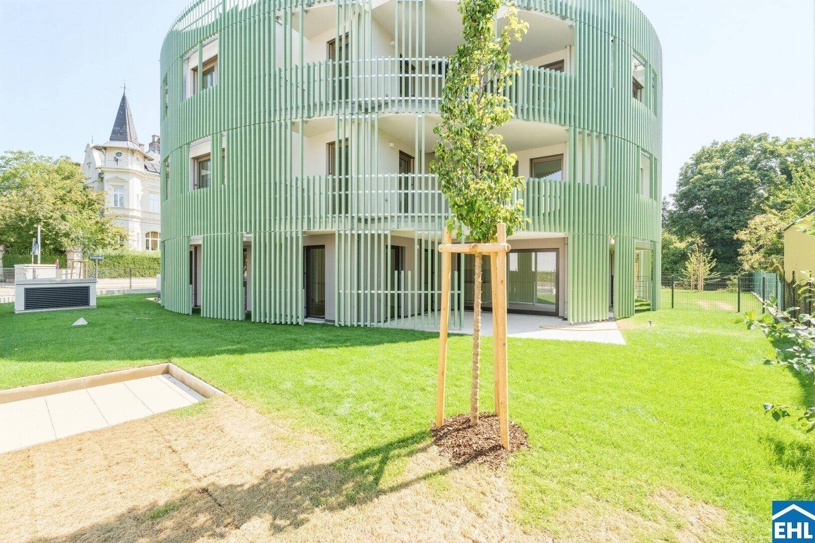 Modernes, rundes Gebäude mit grüner Fassade, umgeben von einer gepflegten Rasenfläche und jungen Bäumen.