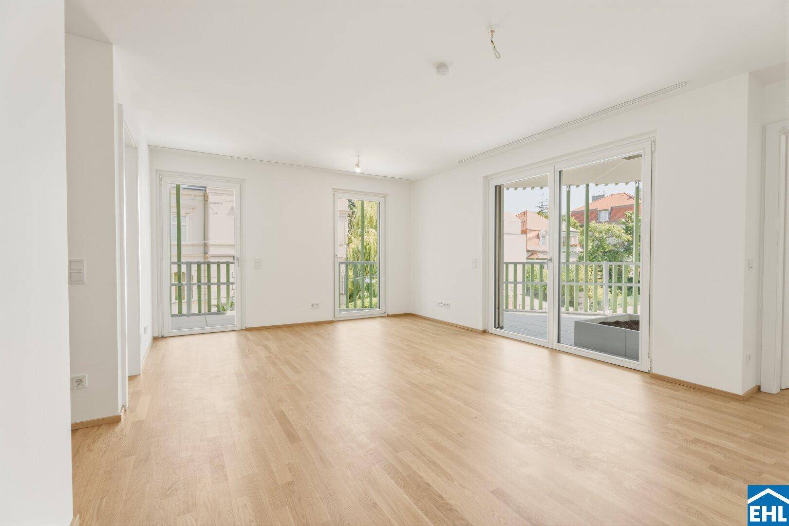 Helles Wohnzimmer mit Parkettboden und Zugang zu mehreren Balkonen, umgeben von Grün.