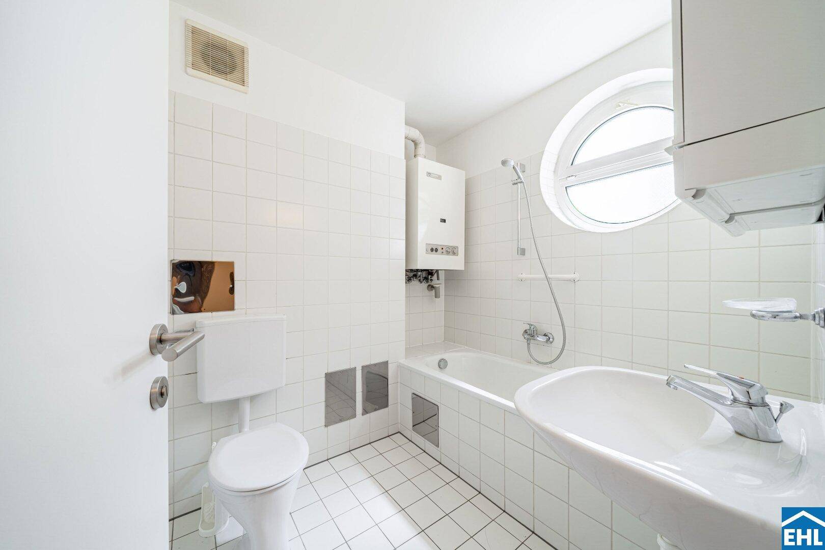 Weiß gefliestes Badezimmer mit Badewanne, WC, Waschbecken und einem markanten runden Fenster.