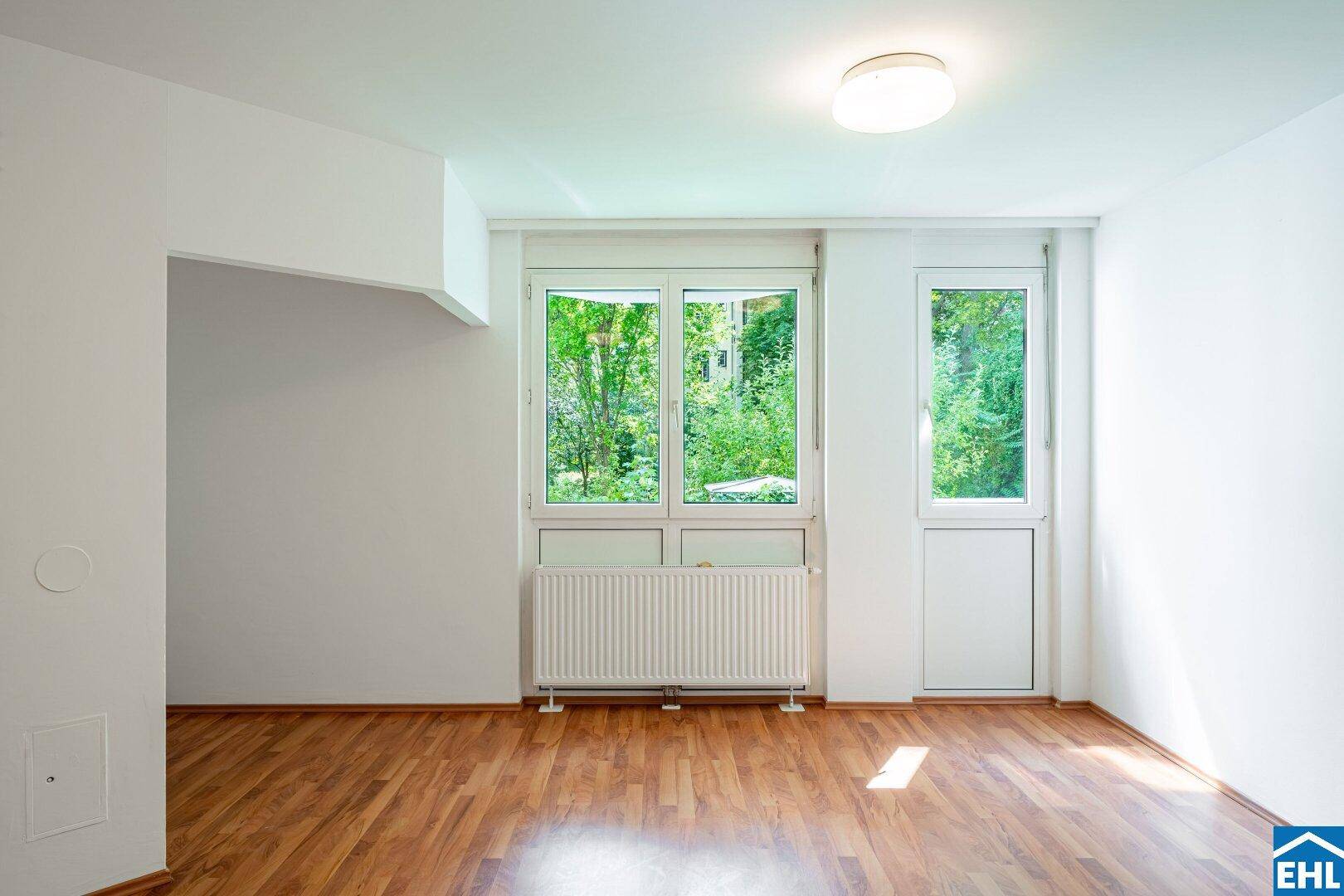 Lichtdurchflutetes Zimmer mit Blick ins Grüne und einem Heizkörper unter den Fenstern.