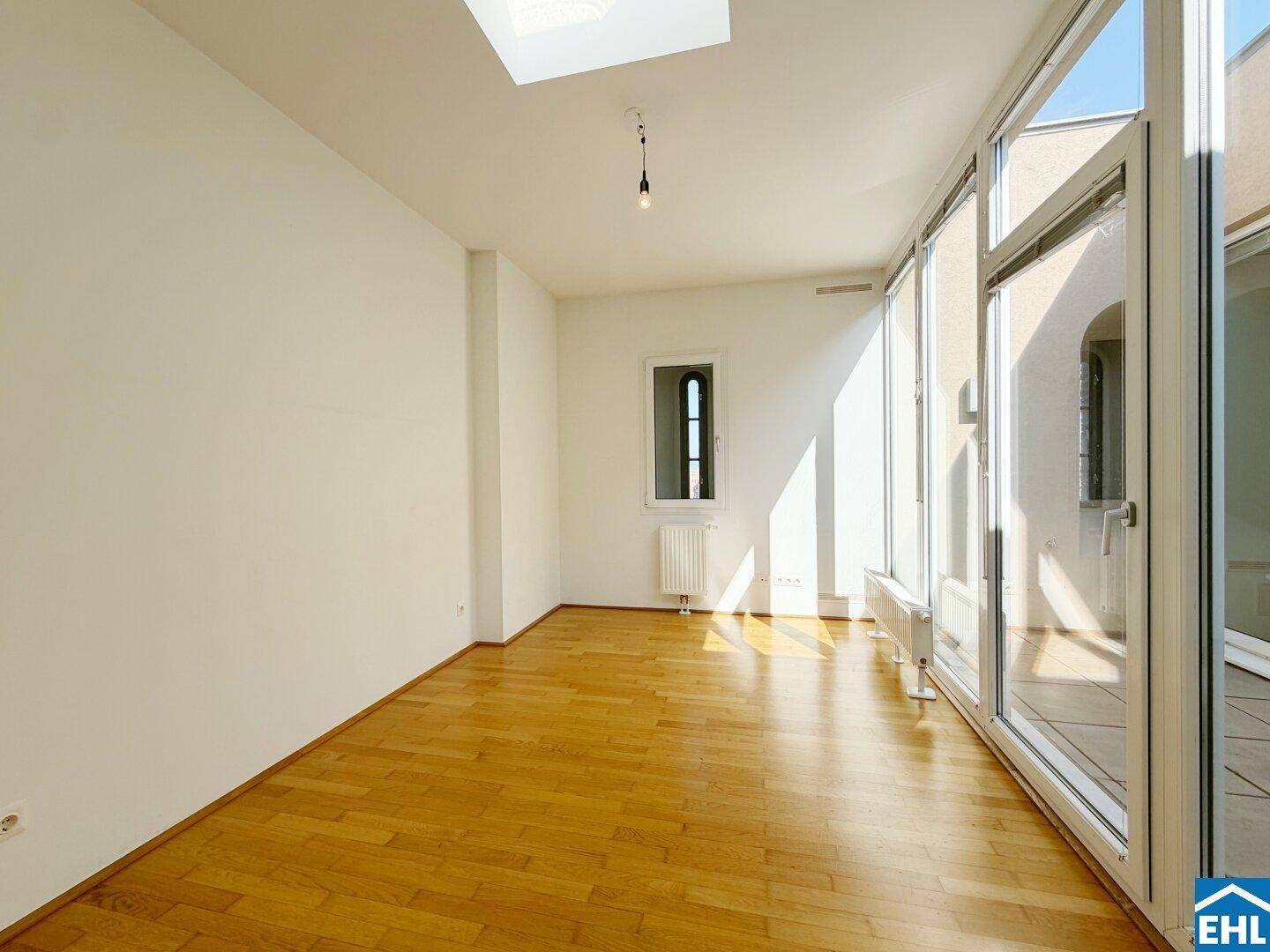 Helles Zimmer mit Parkettboden, hohen Decken und großen Fenstern, die viel Licht hereinlassen.