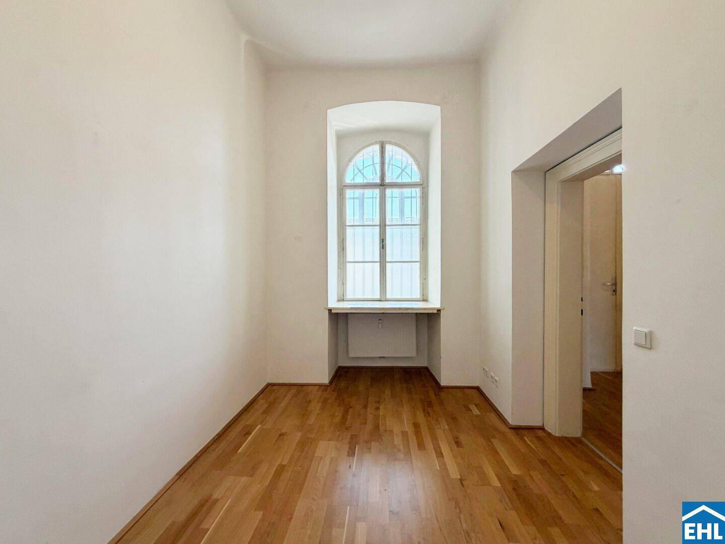 Helles Zimmer mit Holzdielenboden und einem hohen Bogenfenster.