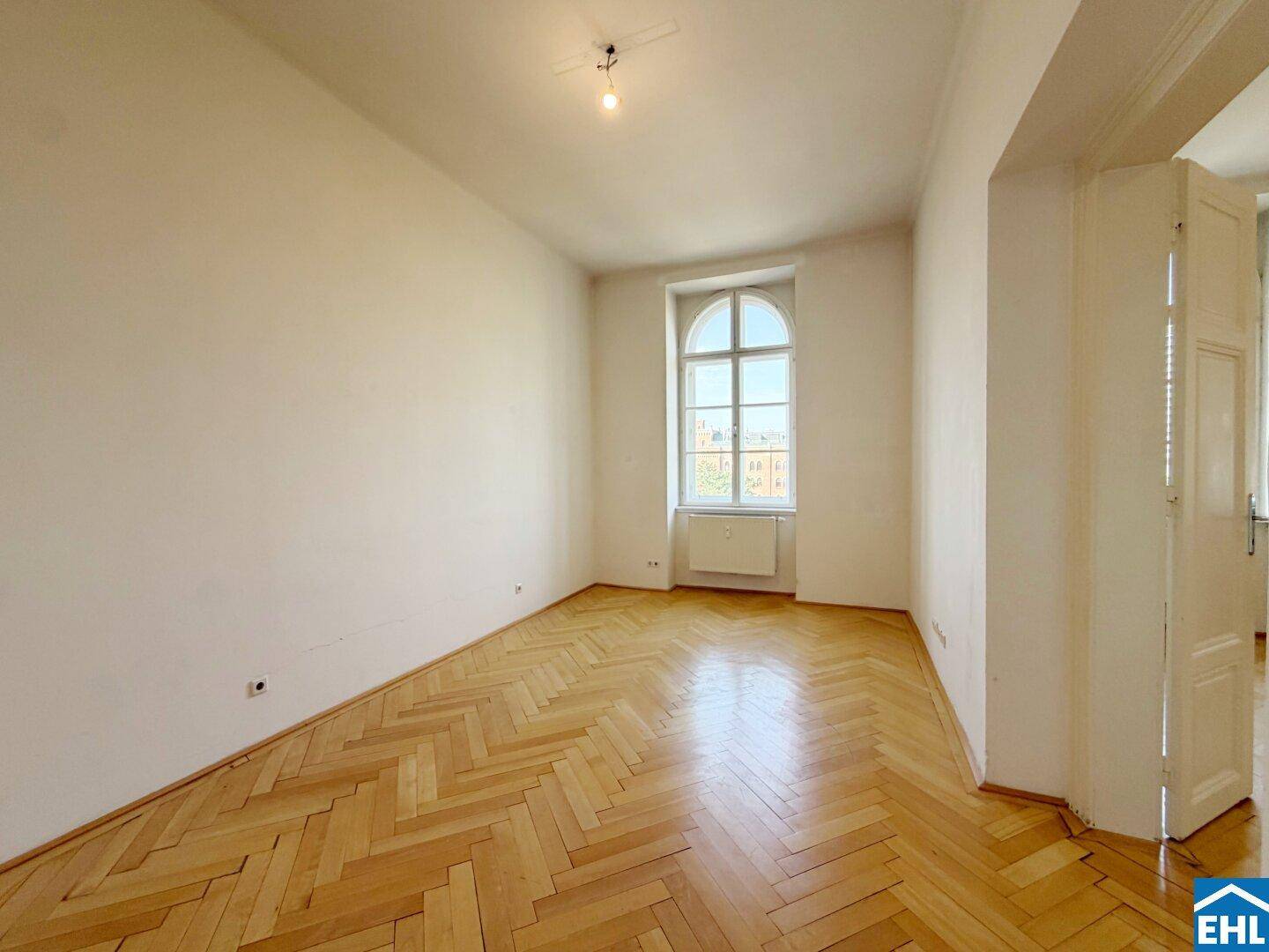 Geräumiges Zimmer mit Fischgrätparkett, hohem Bogenfenster und Doppeltür.