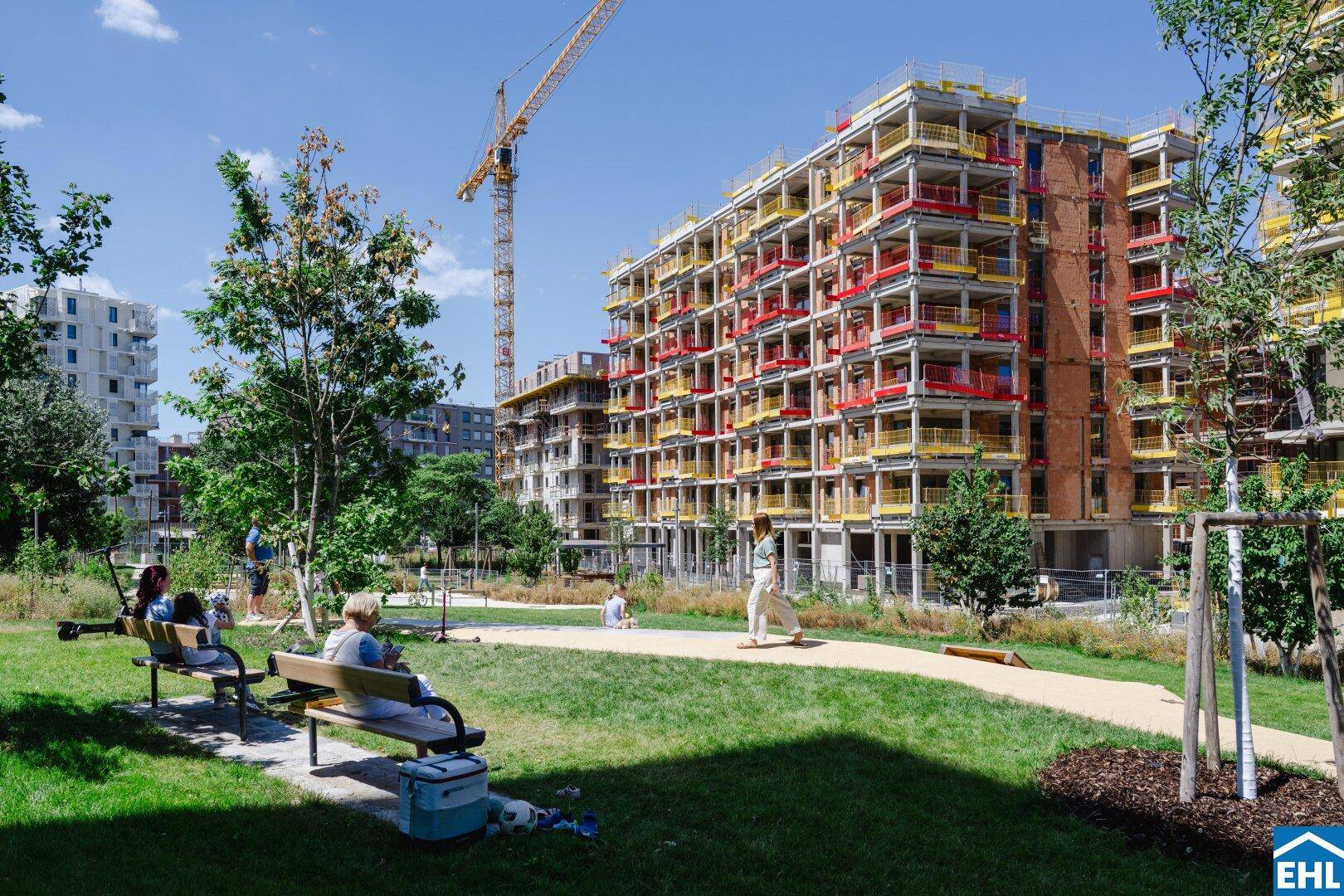 Weitläufige Grünflächen mit Wegen und Sitzgelegenheiten um eine moderne Wohnanlage im Bau.
