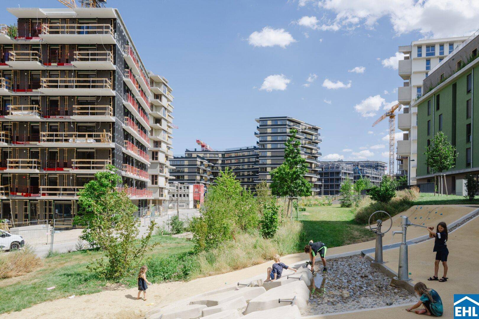 Moderne Wohngebäude umgeben von Grünflächen mit spielenden Kindern an einem Wasserspielplatz.