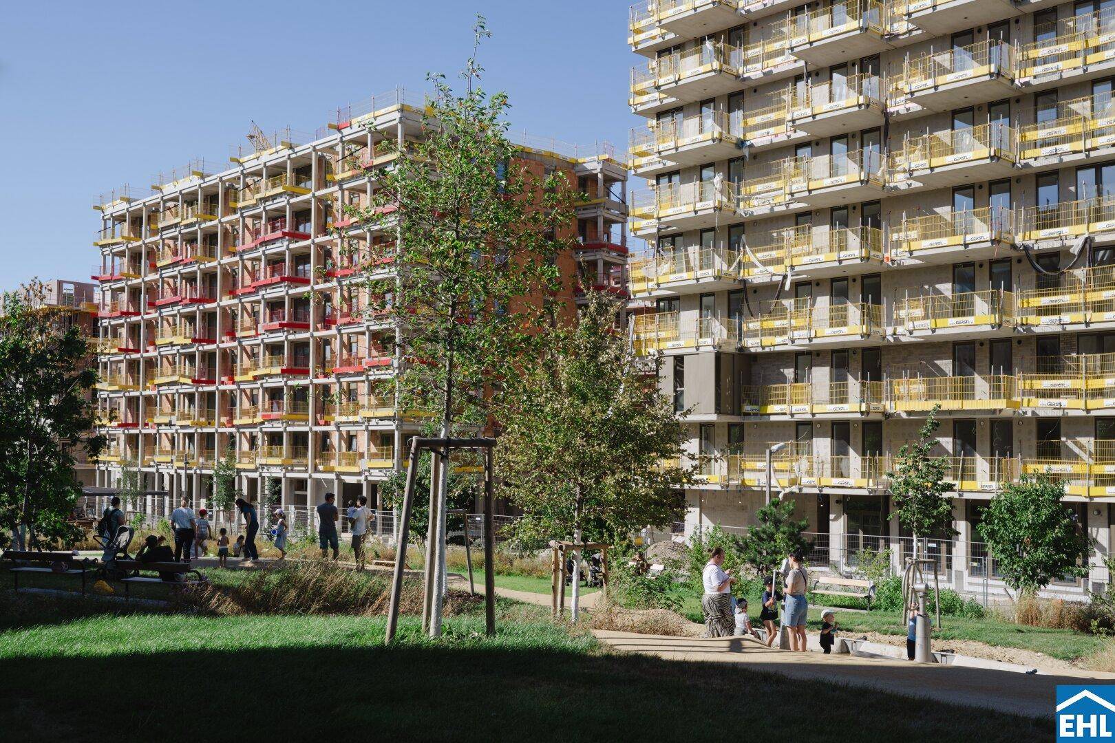 Parkähnliche Grünfläche mit Bäumen und spielenden Menschen, umgeben von im Bau befindlichen Wohngebäuden.