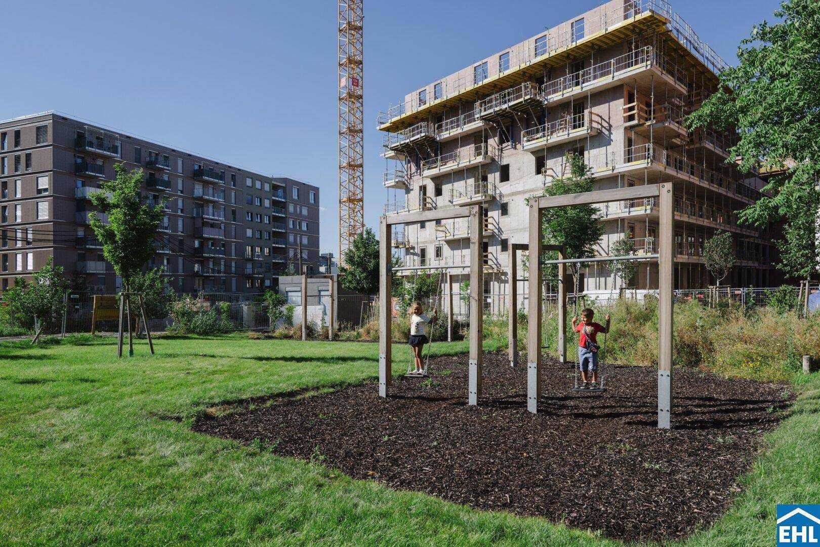 Spielplatz mit Schaukeln auf Rindenmulch, umgeben von Rasen und im Bau befindlichen Wohngebäuden.