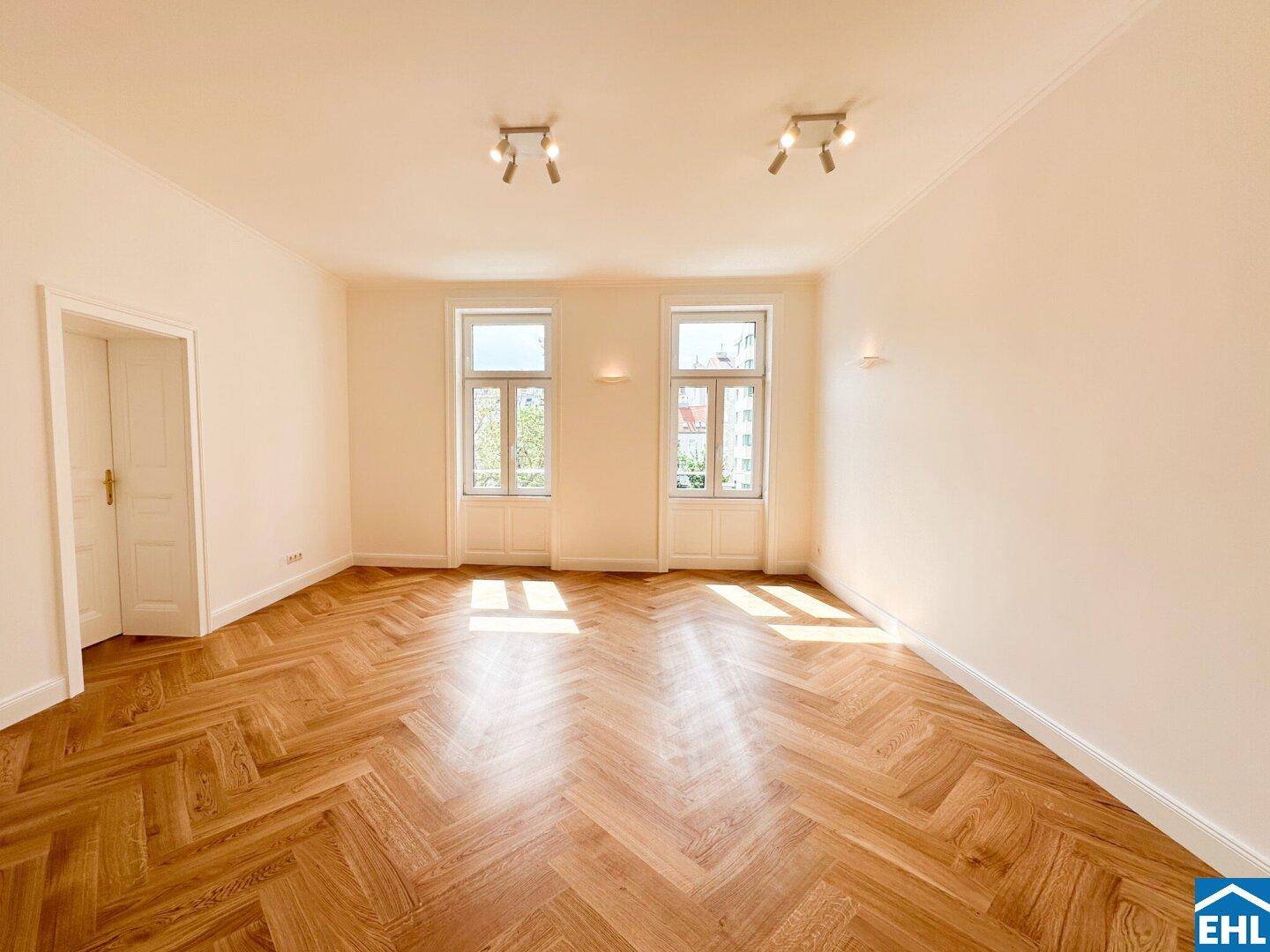Geräumiges Wohnzimmer mit elegantem Fischgrätparkett, hohen Fenstern und viel natürlichem Licht.