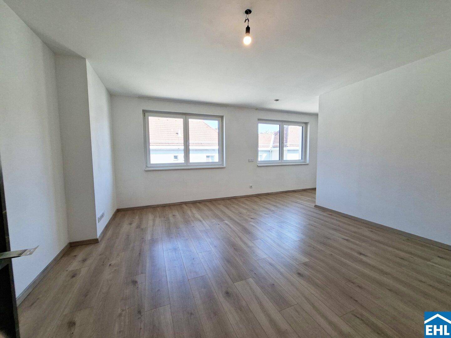 Geräumiger Wohnbereich mit hellem Laminatboden und zwei großen Fenstern.