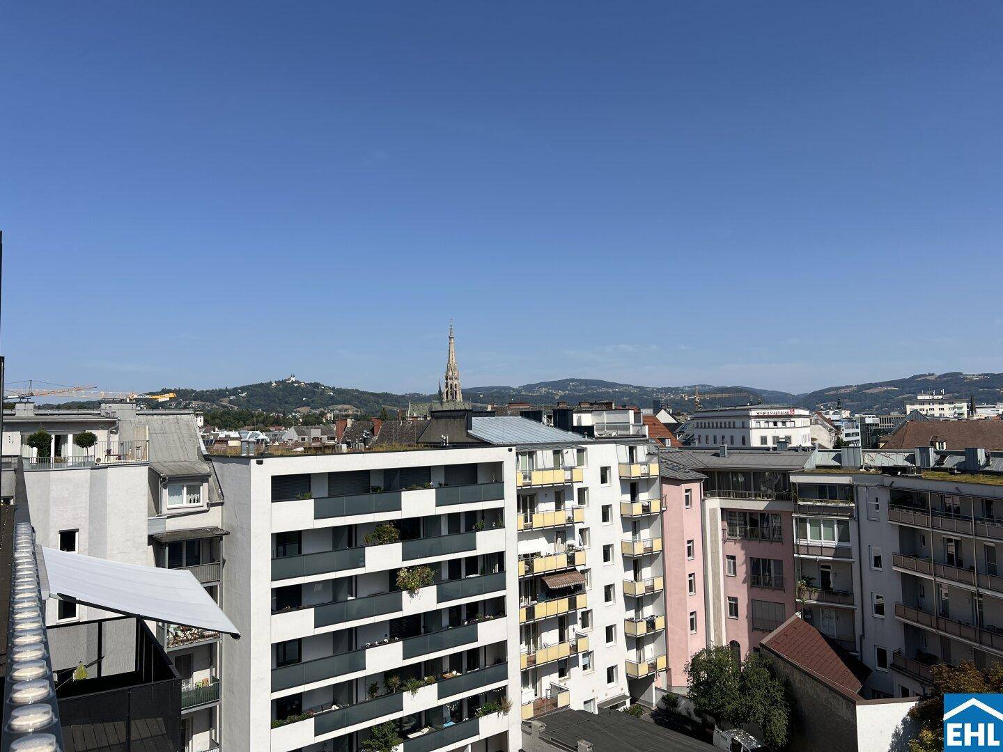 Weitläufiger Stadtblick mit vielen Gebäuden und grünen Hügeln am Horizont unter klarem Himmel.