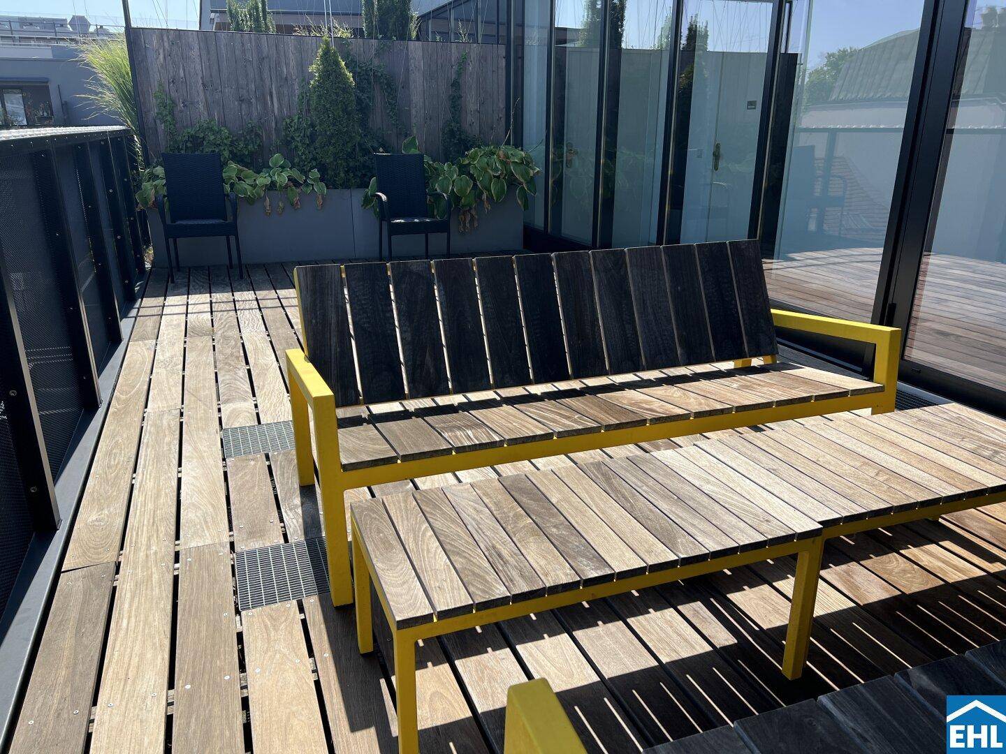 Holzterrasse mit Esstisch und Bänken, umgeben von begrünten Pflanzgefäßen, ideal für Mahlzeiten im Freien.