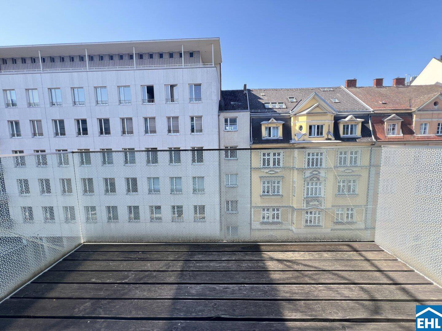 Leerer Balkon mit Holzdeck und Glasgeländer, mit Blick auf die umliegenden Stadtgebäude.