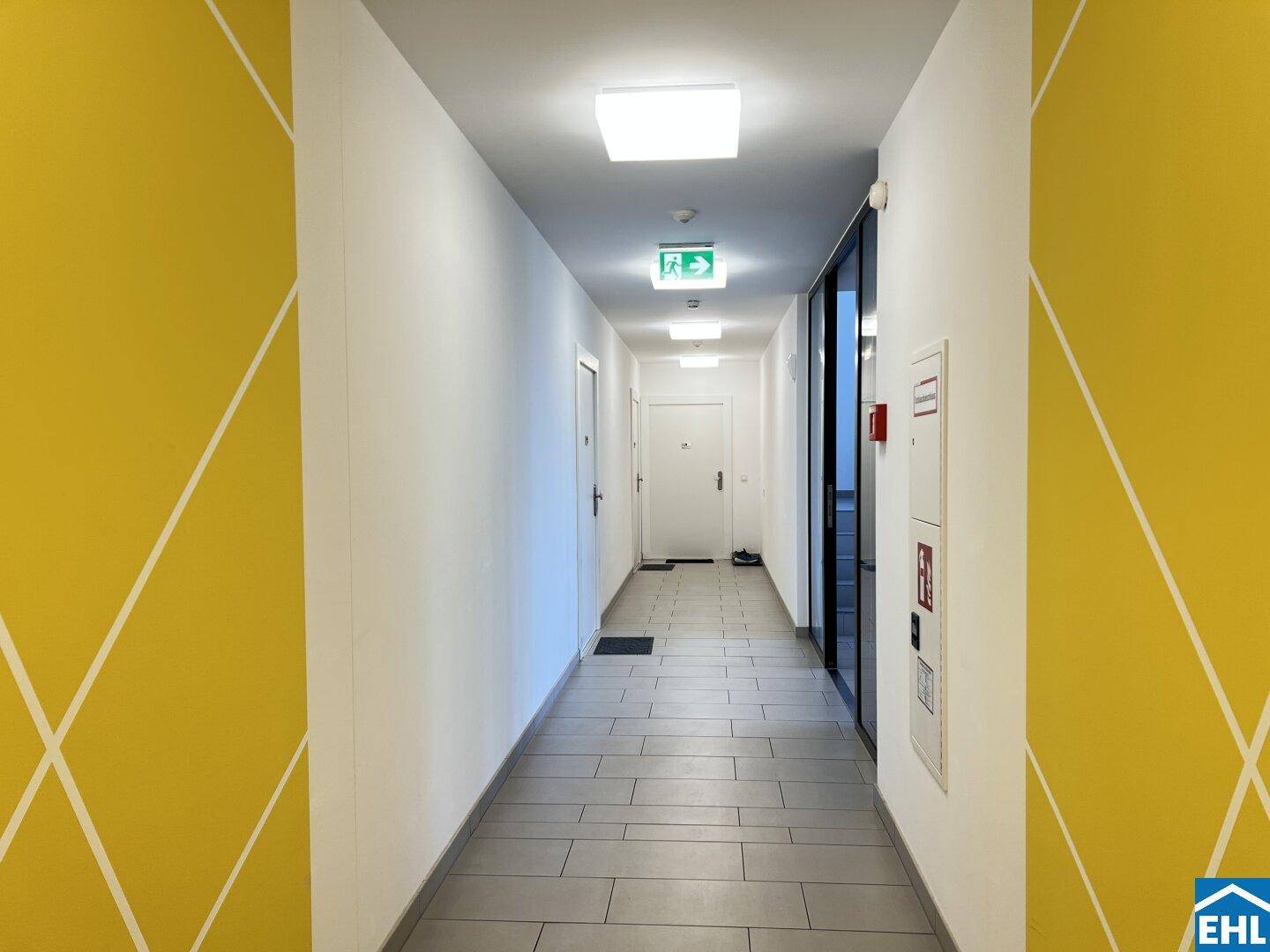 Langer, heller Flur mit Fliesenboden und gelben Akzenten an den Wänden, in einem modernen Gebäude.