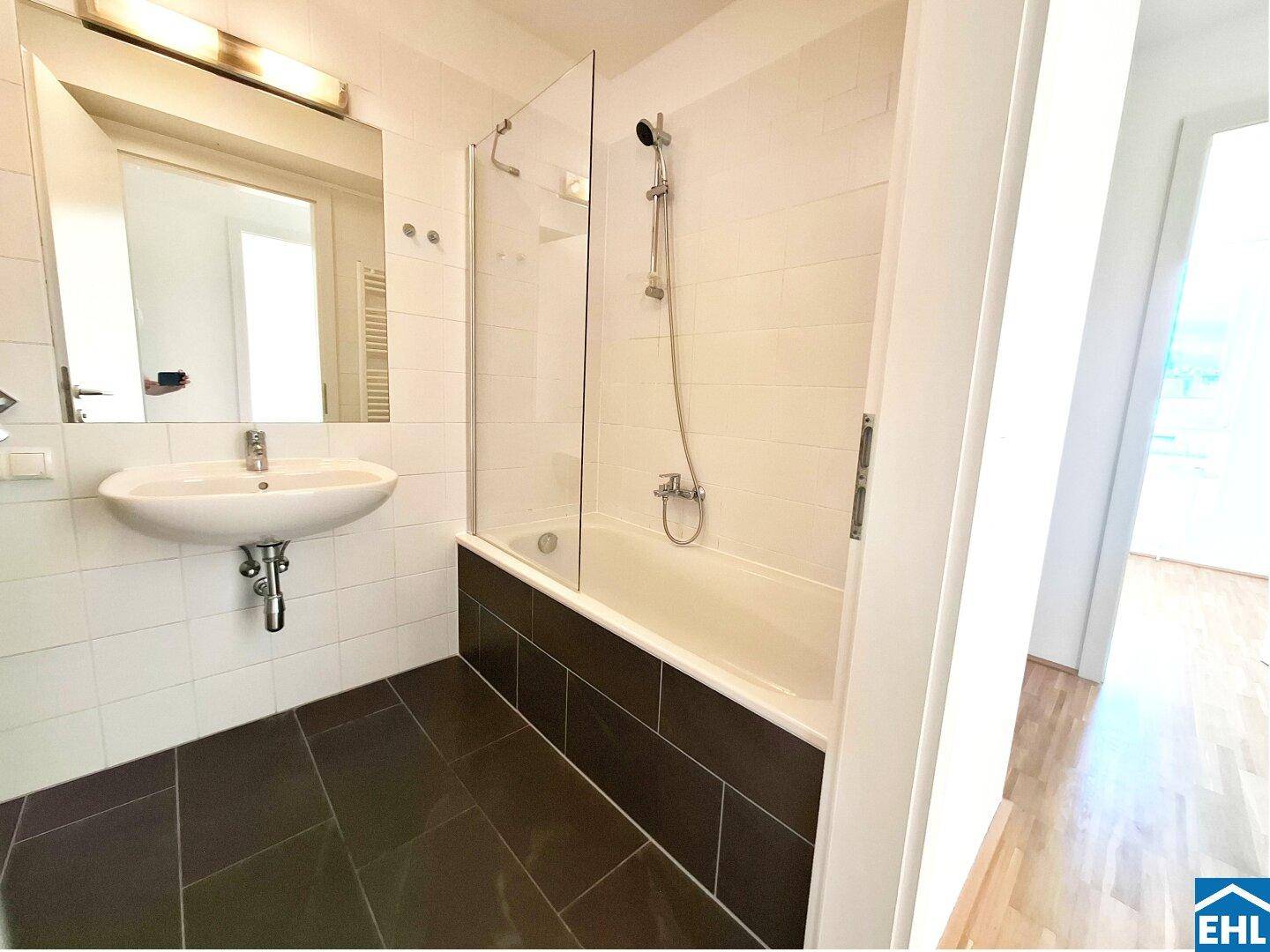 Helles Badezimmer mit weißen Wandfliesen, dunklem Fliesenboden und Badewanne mit Glasduschabtrennung.