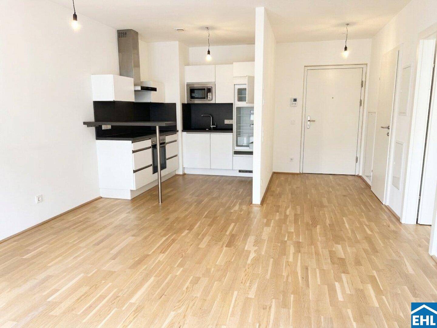 Geräumiger, leerer Wohnbereich mit offener Küchenzeile und hellem Parkettboden in einem Neubau.