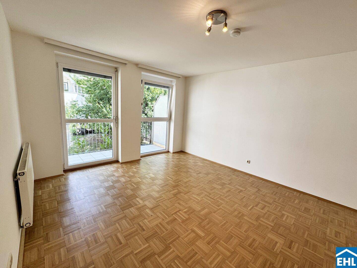 Lichtdurchfluteter Wohnraum mit Parkettboden und zwei Fenstertüren, die viel Tageslicht hereinlassen.