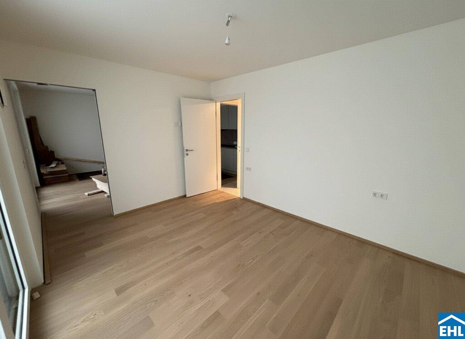 Leerer Raum mit hellem Parkettboden und Zugang zu weiteren Zimmern und einem Fenster.