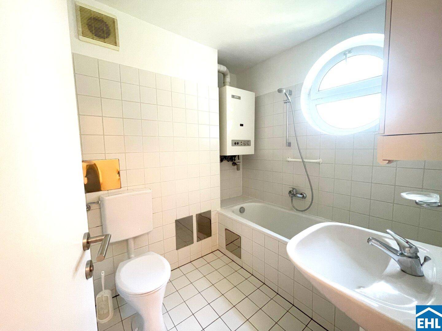 Weiß gefliestes Badezimmer mit Badewanne, WC, Waschbecken und einem markanten runden Fenster.