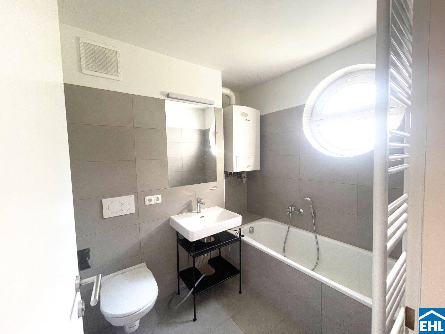 Modernes Badezimmer mit grauen Fliesen, Badewanne, runden Fenster und Handtuchheizkörper.