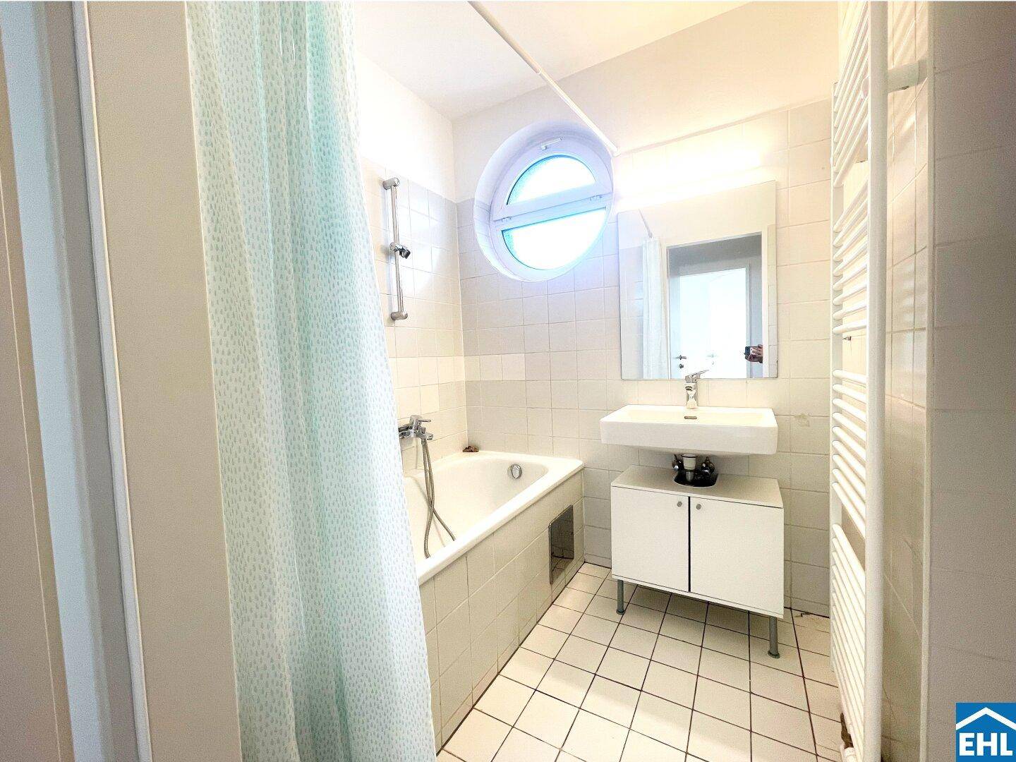 Helles Badezimmer mit Badewanne, Waschbecken und Handtuchheizkörper, ausgestattet mit Rundfenster.