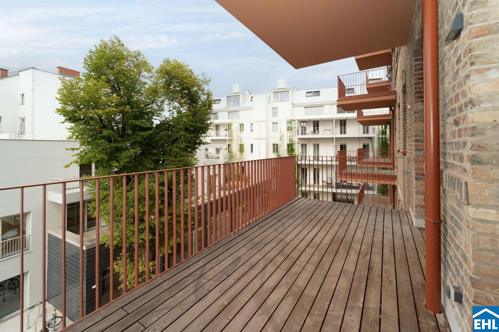 Geräumiger Balkon mit Holzboden und Metallgeländer, umgeben von modernen Gebäuden und Bäumen.