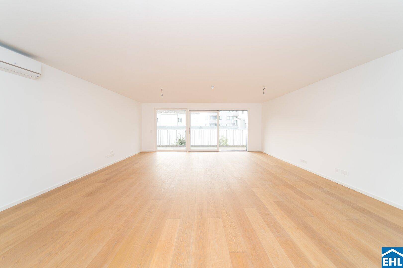 Helles, leeres Zimmer mit Holzboden und Zugang zu einem Balkon, ausgestattet mit Klimaanlage.