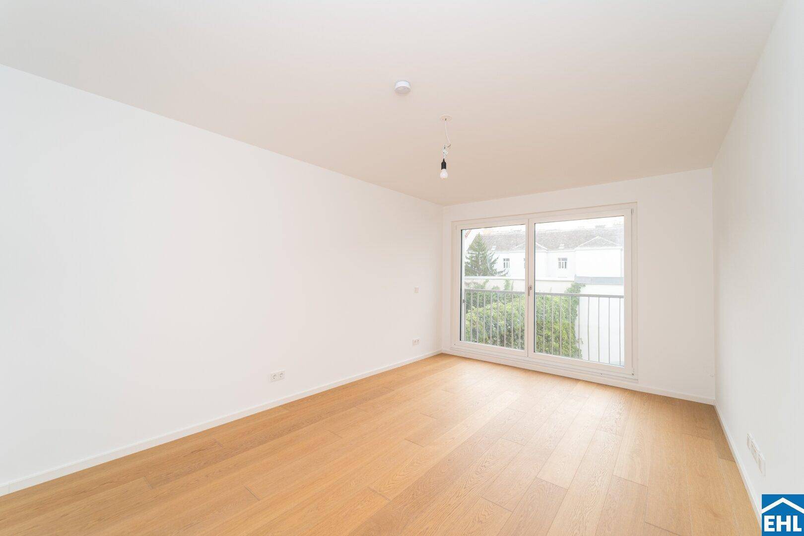 Leeres Zimmer mit Holzboden und Blick ins Grüne durch die Balkontür.