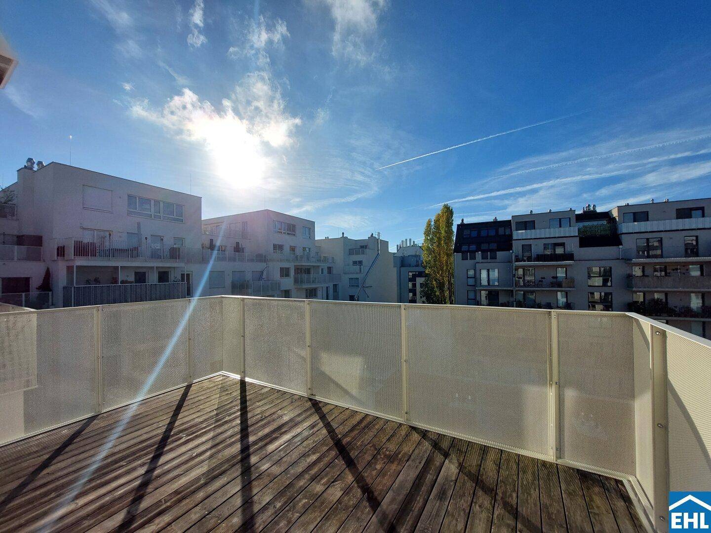 Sonniger Balkon mit Holzboden und Blick auf umliegende Wohngebäude unter blauem Himmel.