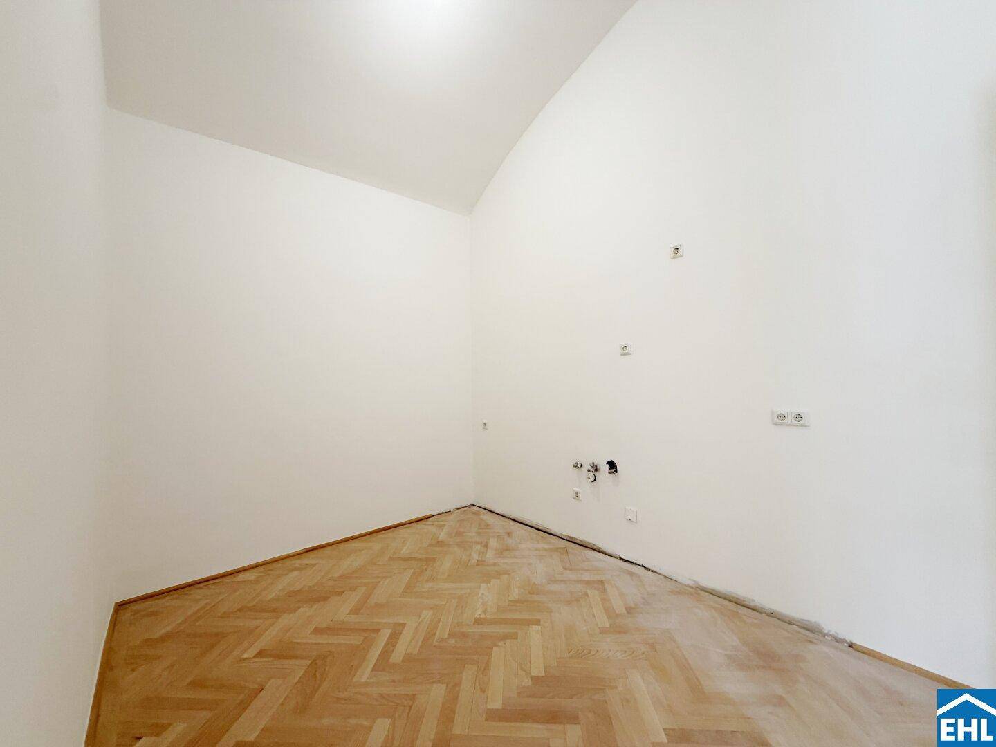 Leerer Raum mit schrägen Wänden, Fischgrätparkett und Anschlüssen für eine mögliche Küchenzeile.