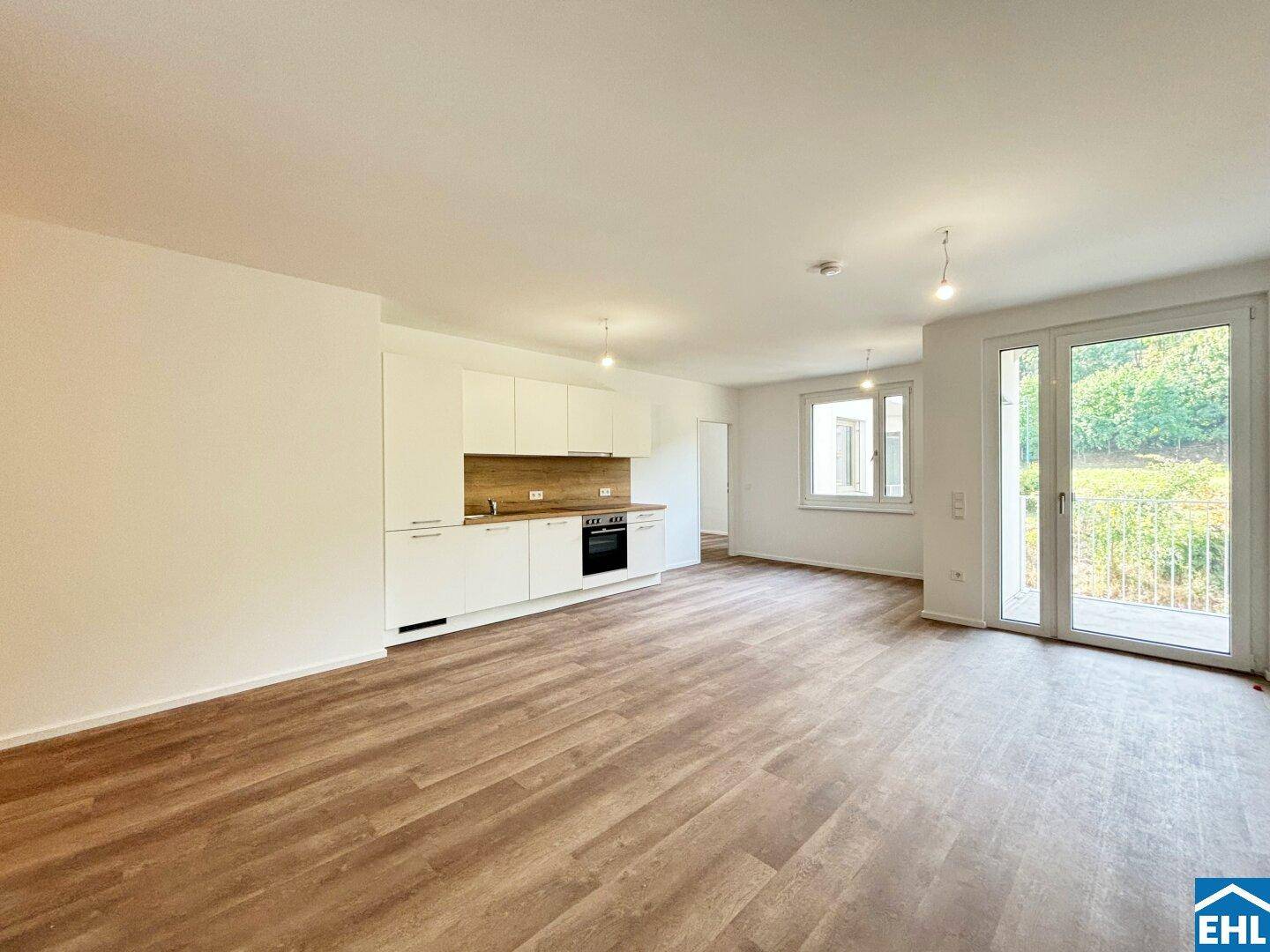 Heller offener Wohnbereich mit integrierter Küchenzeile, Laminatboden und Zugang zum Balkon.
