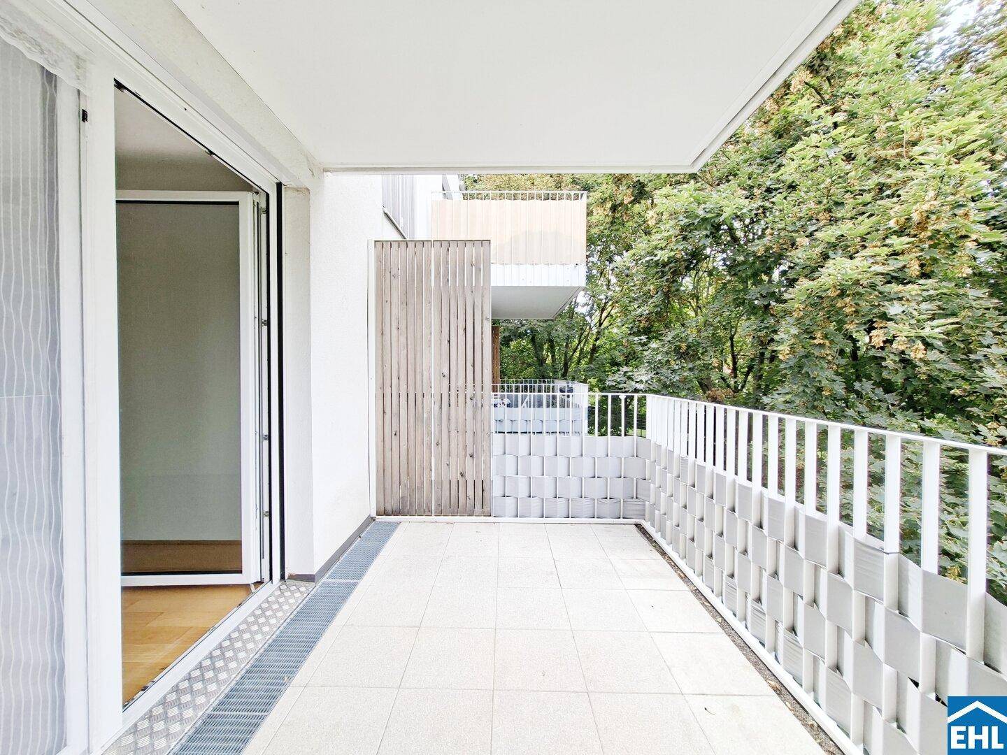 Heller Balkon mit moderner Brüstung und Zugang zum Innenbereich der Wohnung, umgeben von Bäumen.