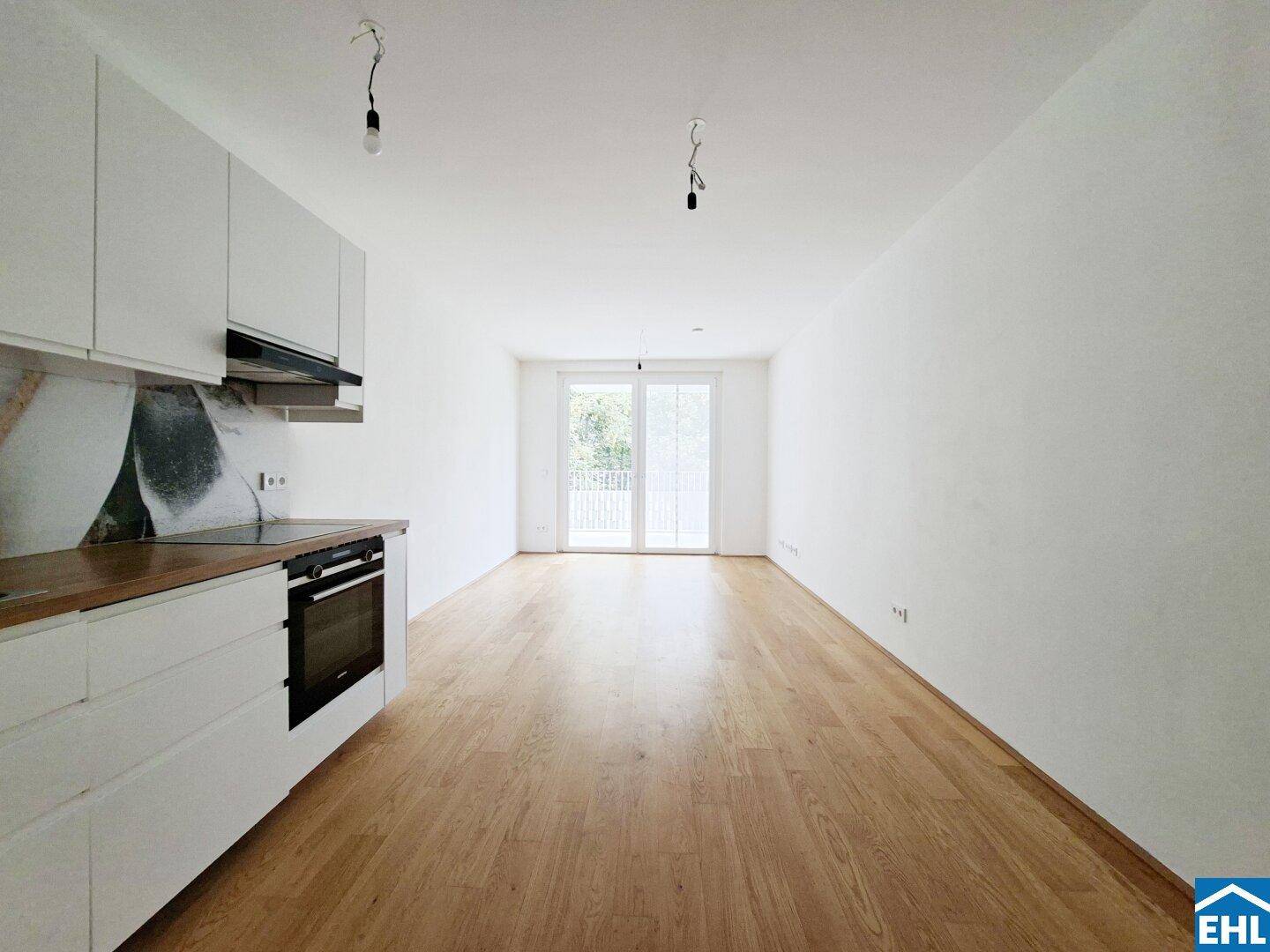 Offener Wohnbereich mit integrierter weißer Küche, Holzboden und Zugang zum Balkon.