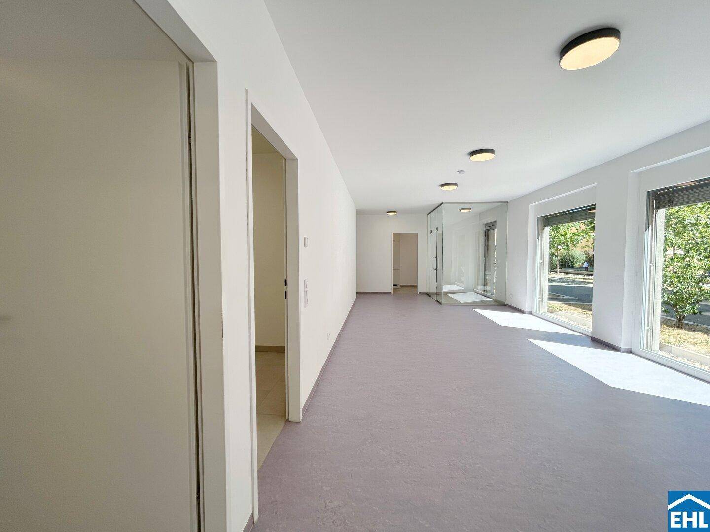 Langer, heller Raum mit mehreren Türen und großen Fenstern, die den Raum mit Licht füllen.
