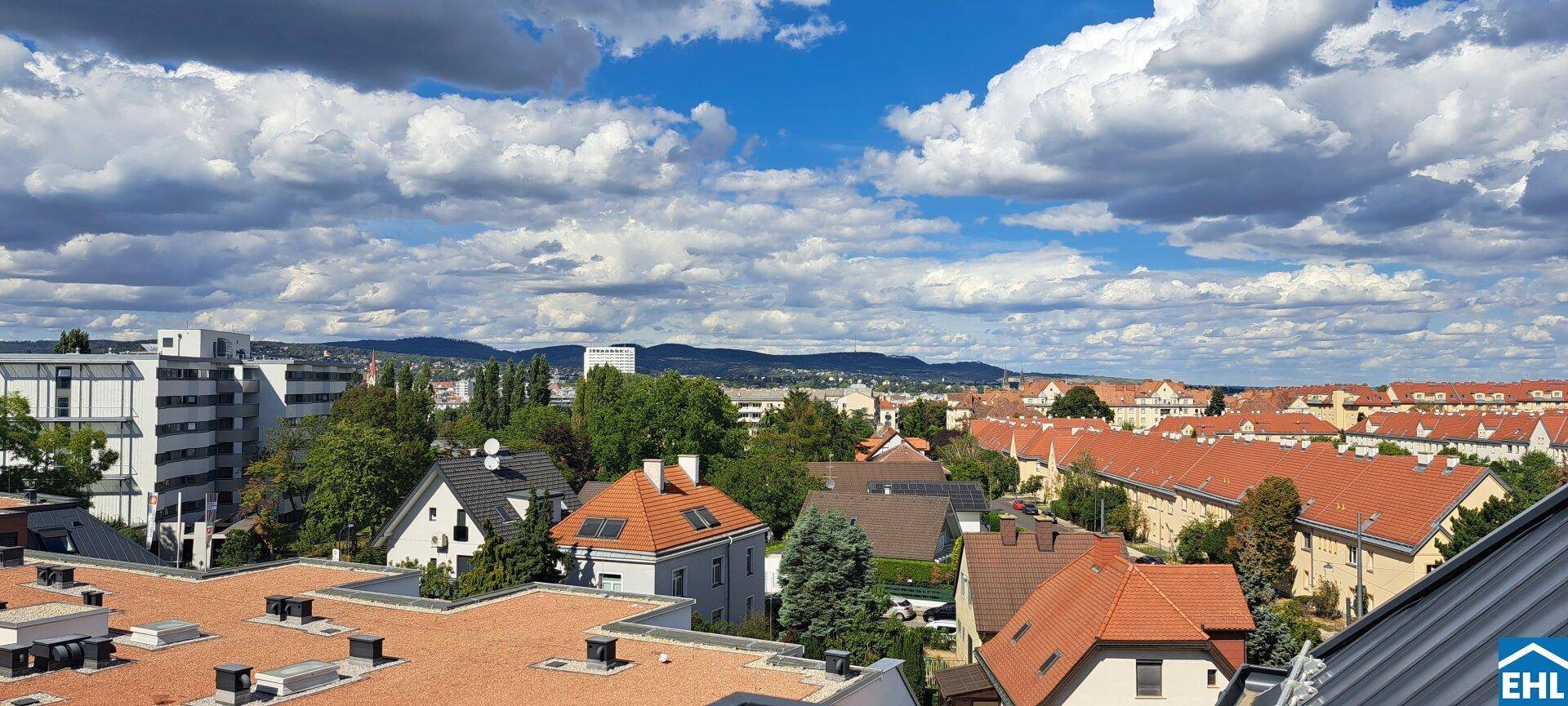 Weitblick über die städtische Bebauung mit roten Dächern und bewaldeten Hügeln am Horizont.