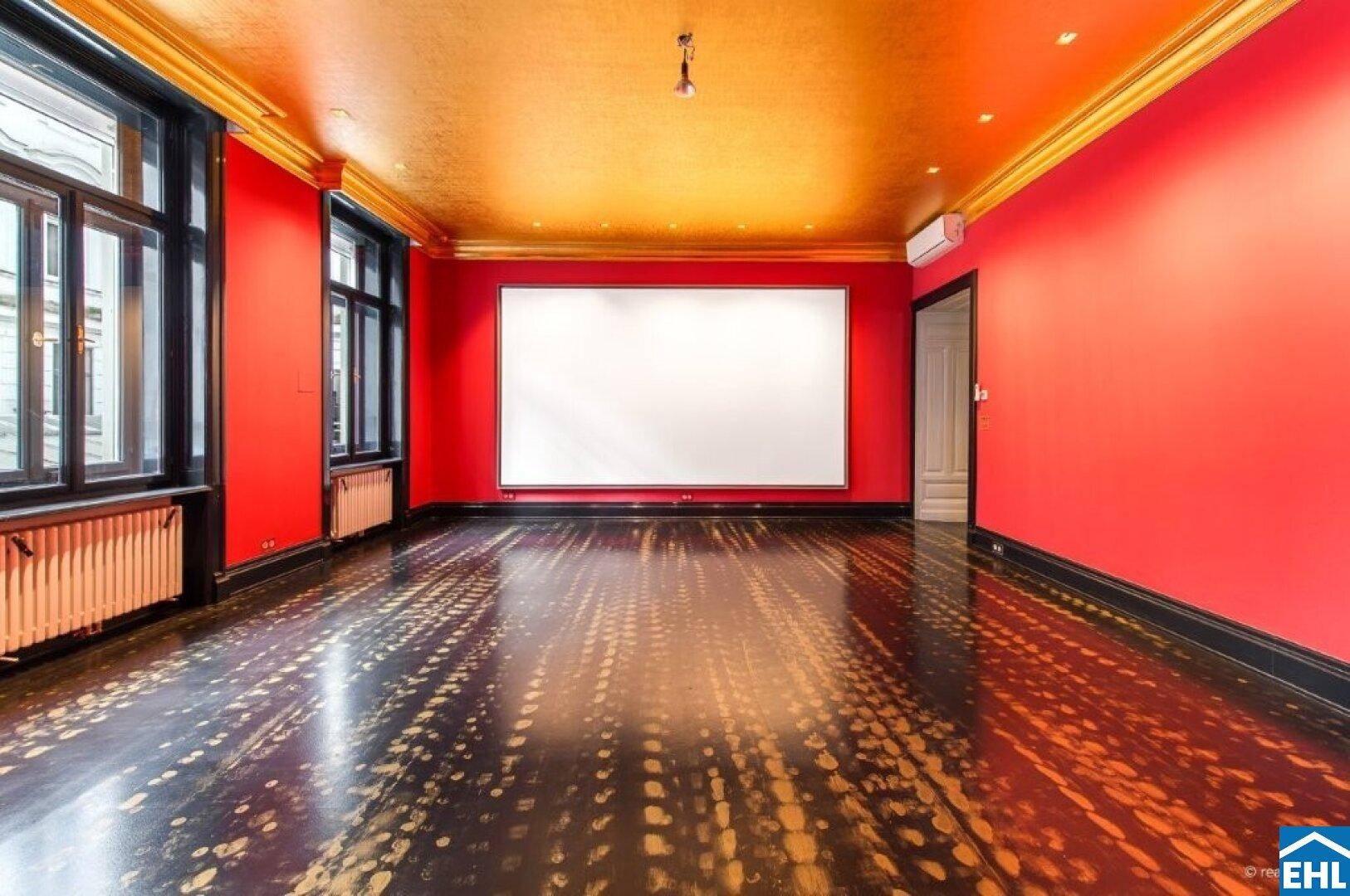 Großer Raum mit roter Wandfarbe, goldener Decke und einem Projektionsbildschirm.