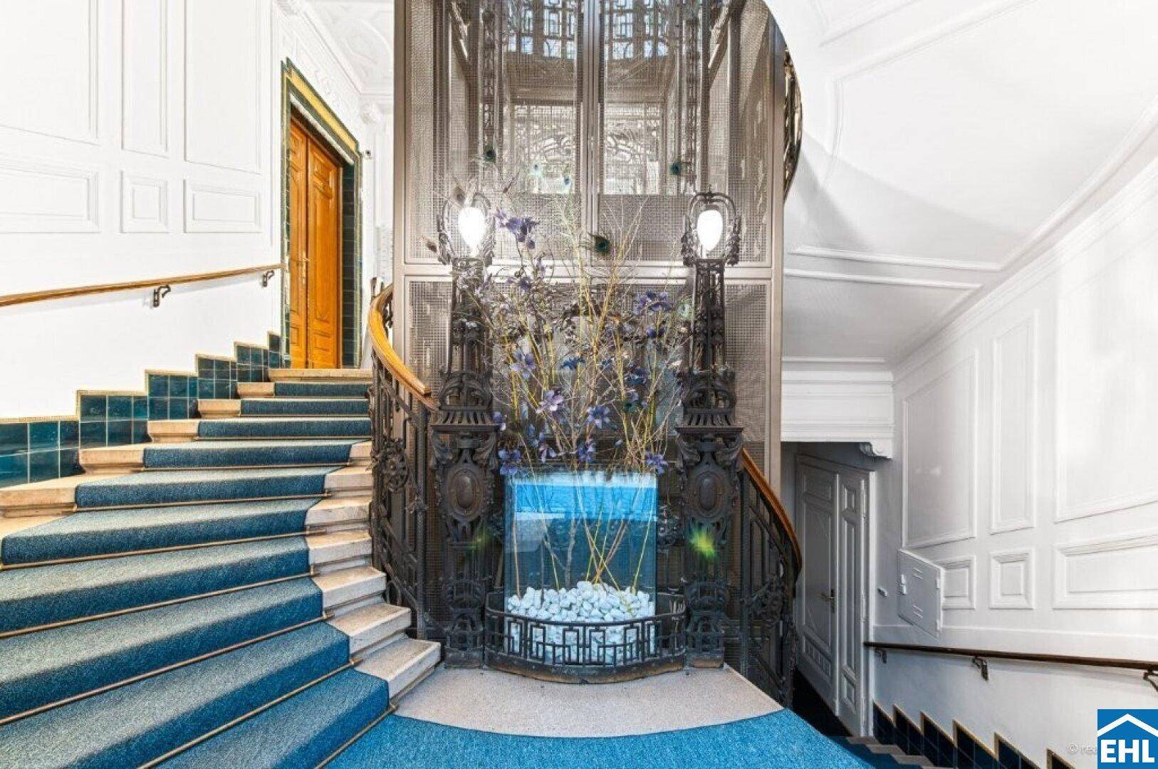 Elegantes Treppenhaus mit blauem Teppich, kunstvollem Geländer und einem historischen Aufzug.