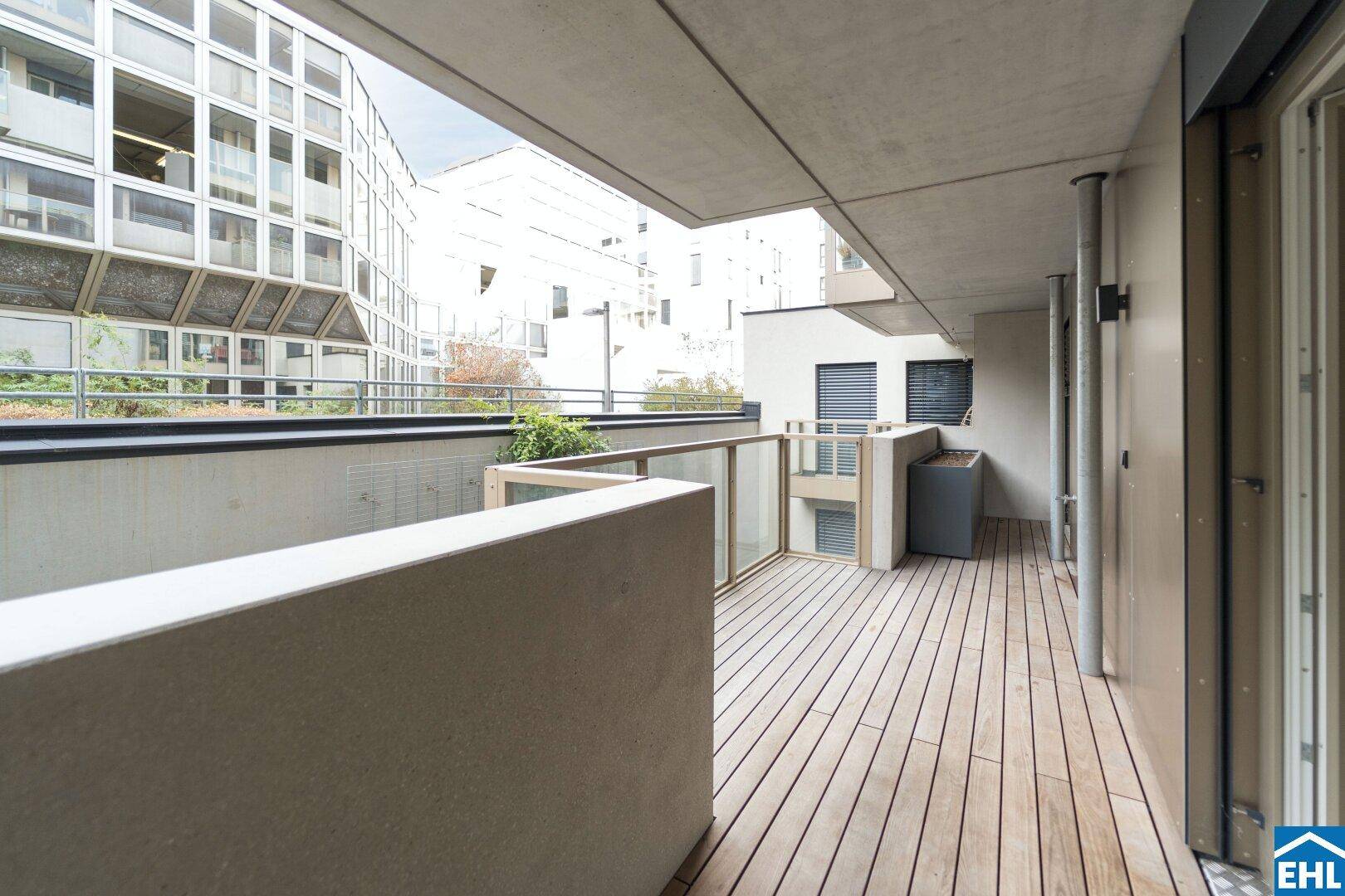 Geräumiger Balkon mit Holzboden und Glasgeländer, umgeben von moderner Architektur.