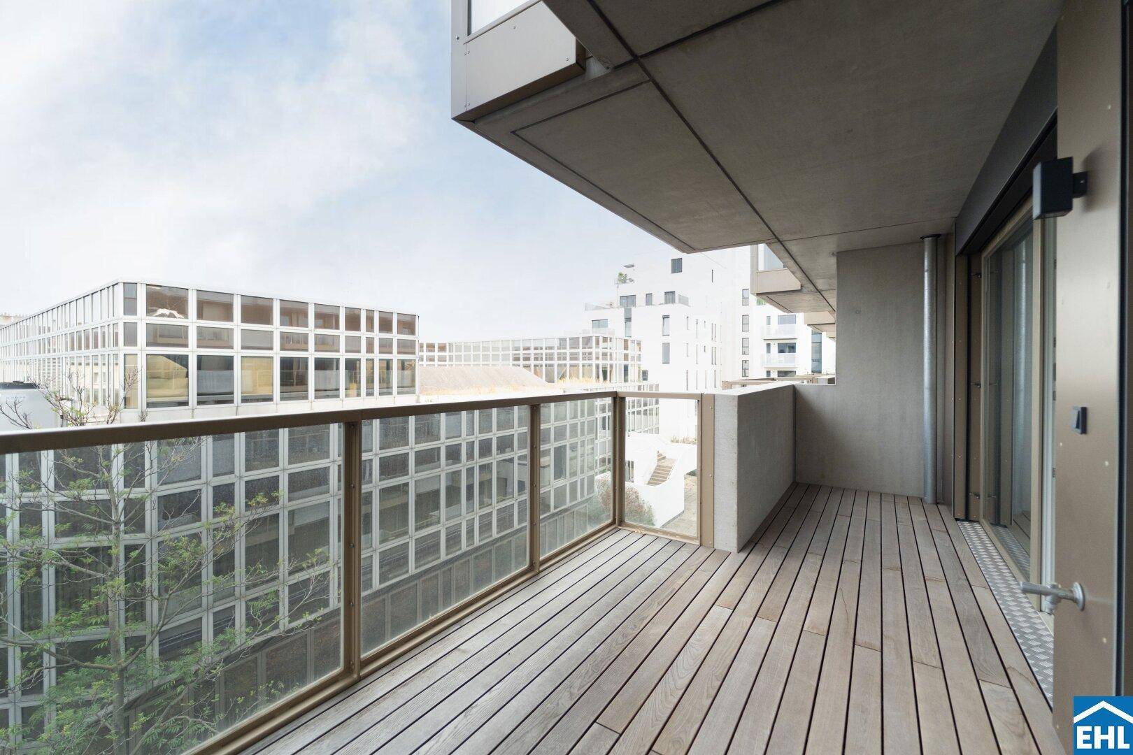 Breiter Balkon mit Holzboden und Glasgeländer, umgeben von moderner Stadtarchitektur.