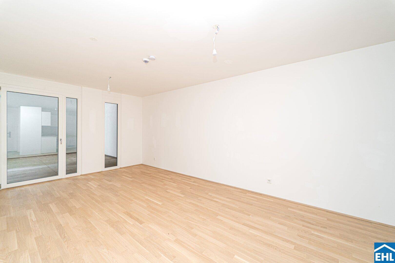 Geräumiges, helles Zimmer mit Holzboden und großen Fenstern, die eine Küche spiegeln.