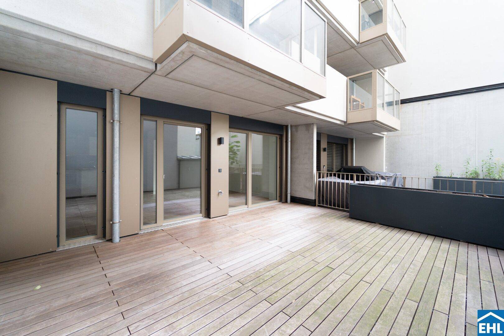 Moderne Gebäudefassade mit großen Glasfronten und einer weitläufigen Holzterrasse im Erdgeschoss.