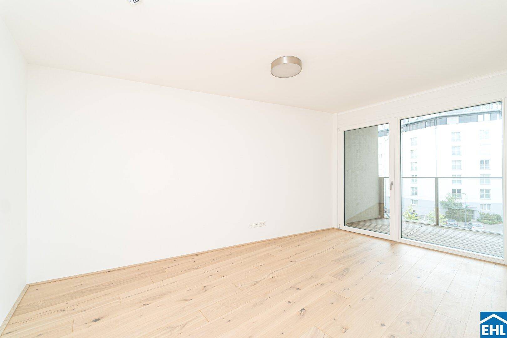 Modernes, helles Zimmer mit Parkettboden und großem Fenster, das den Zugang zum Balkon ermöglicht.