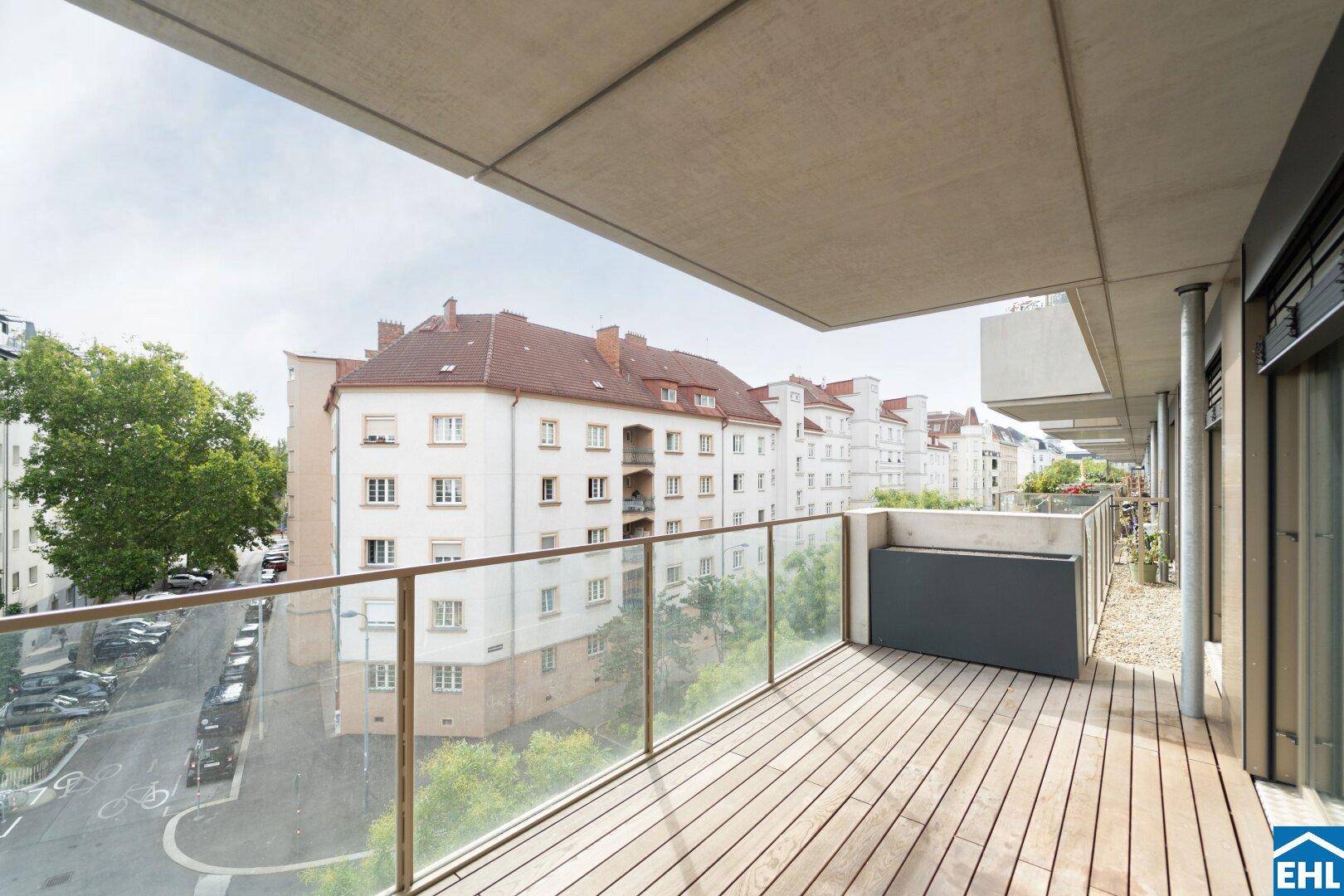 Breiter Balkon mit Holzdielen, Glasbrüstung und Blick auf die umliegenden Gebäude.
