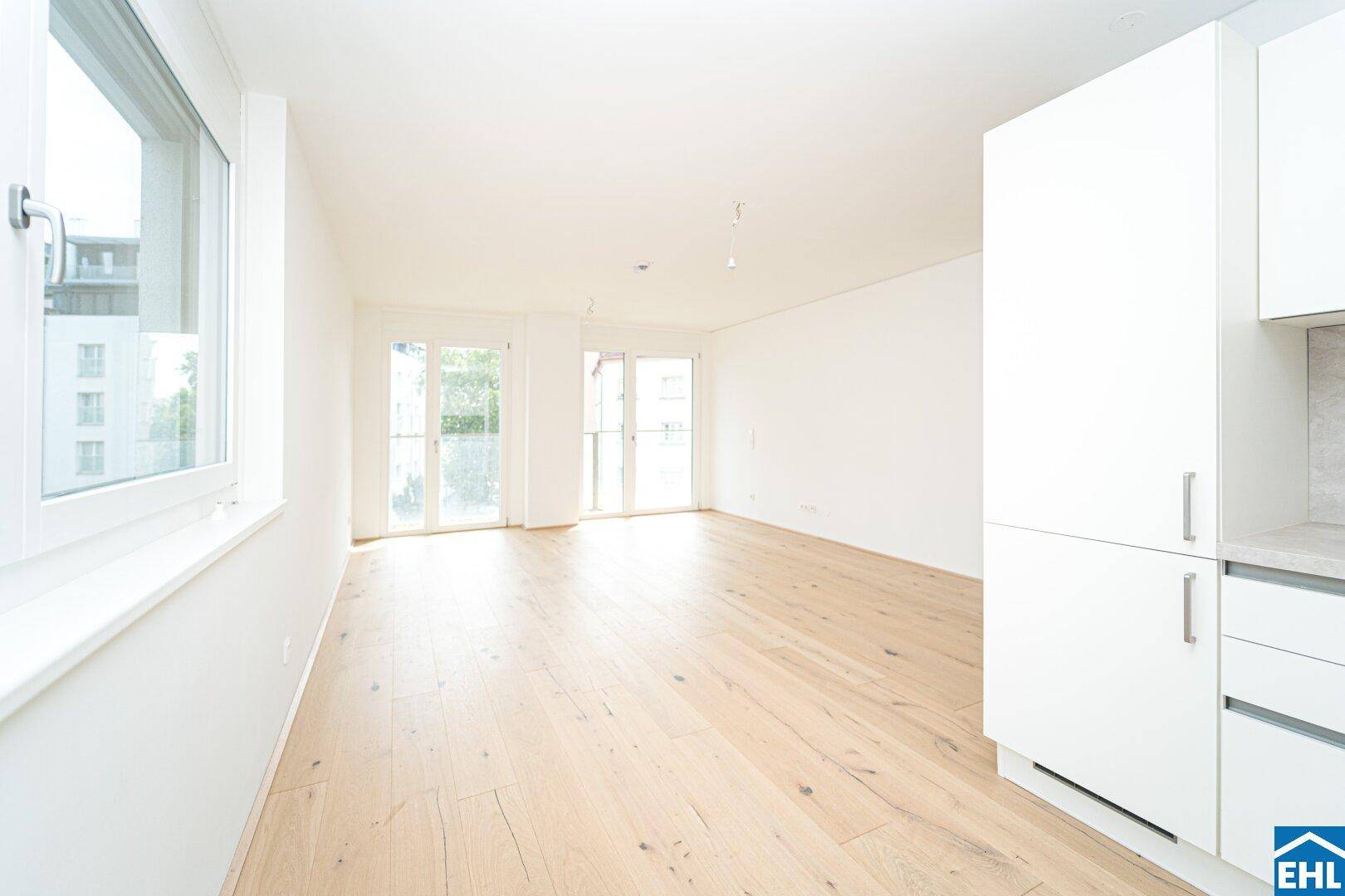Offener Wohnbereich mit Holzboden, großen Fenstern und Zugang zum Balkon.