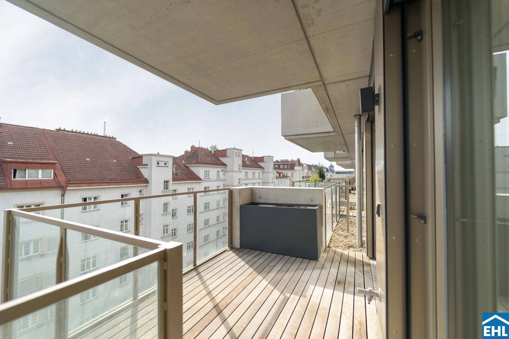 Geräumiger Balkon mit Holzdeck, Glasgeländer und Blick auf die umliegenden Gebäude der Stadt.