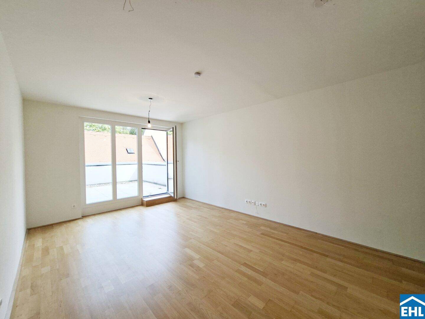 Heller, leerer Raum mit Holzboden und großem Fenster, das viel Tageslicht hereinlässt.