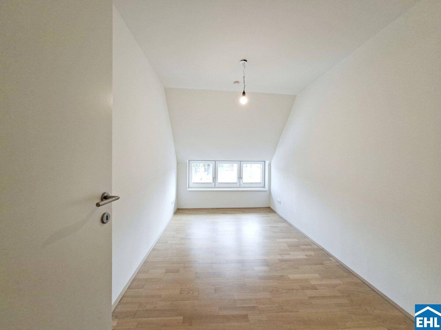 Helles Zimmer mit Dachschrägen, Holzfußboden und einem Fenster mit Ausblick.