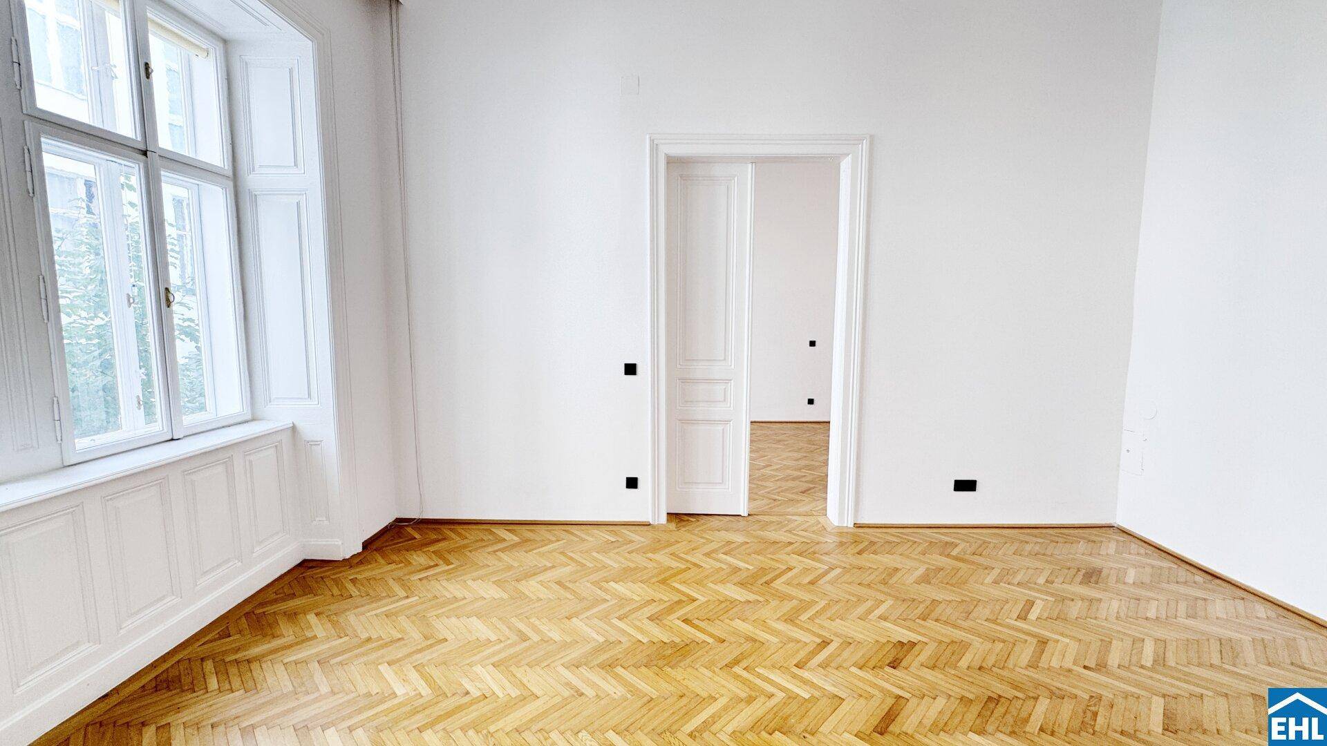 Geräumiges, leeres Zimmer mit Fischgrätparkett, hohen Fenstern und weiß gestrichenen Wänden.