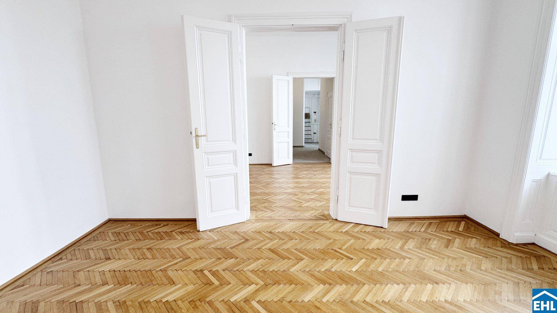 Geräumiges, leeres Zimmer mit Fischgrätparkett und weit geöffneten Doppeltüren zu einem weiteren Raum.