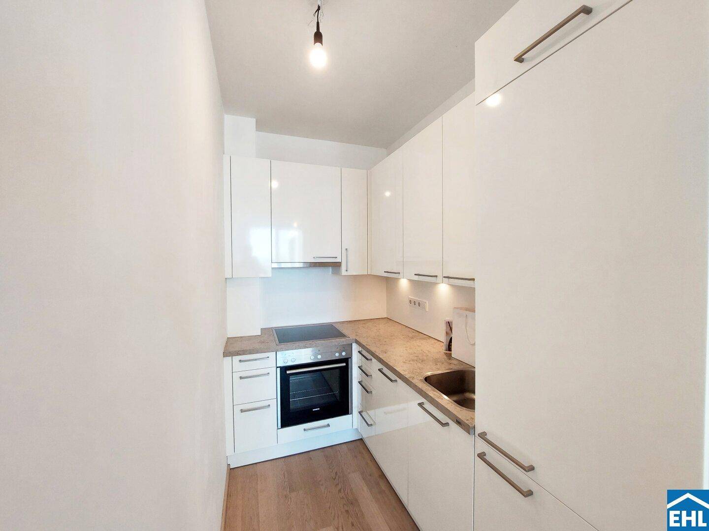 Moderne, kompakte Küche mit weißen Hochglanzfronten, integriertem Backofen und heller Arbeitsplatte.