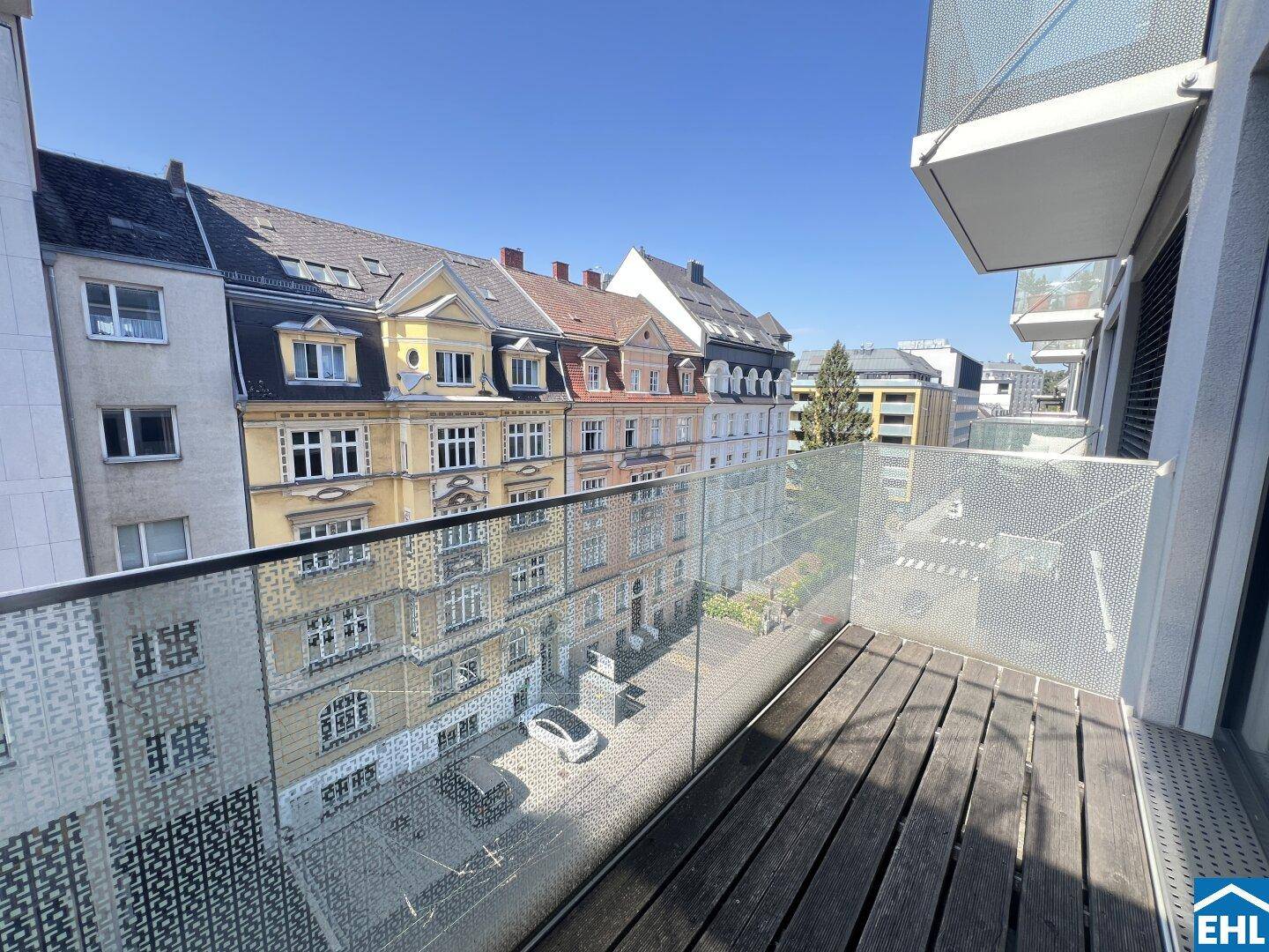 Moderner Balkon mit Holzboden und Glasgeländer, bietet einen sonnigen Blick auf die Stadt und historische Gebäude.